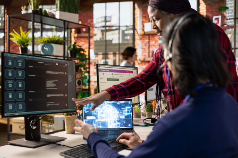 Two professionals collaborating in a modern office, discussing website development strategies, with multiple screens displaying tech-related content and data analytics.